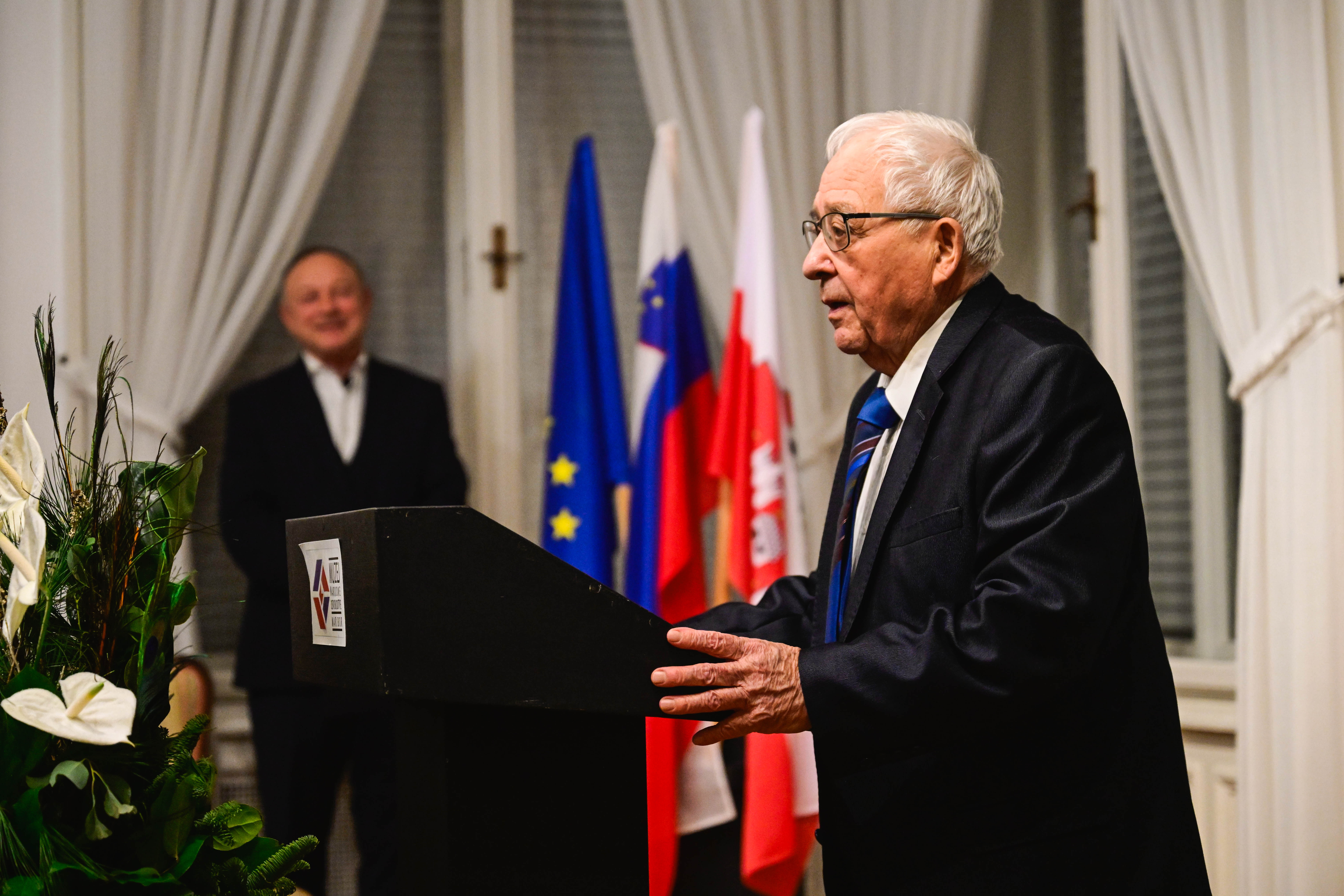 Person at lectern with flags in the background, in a formal setting.