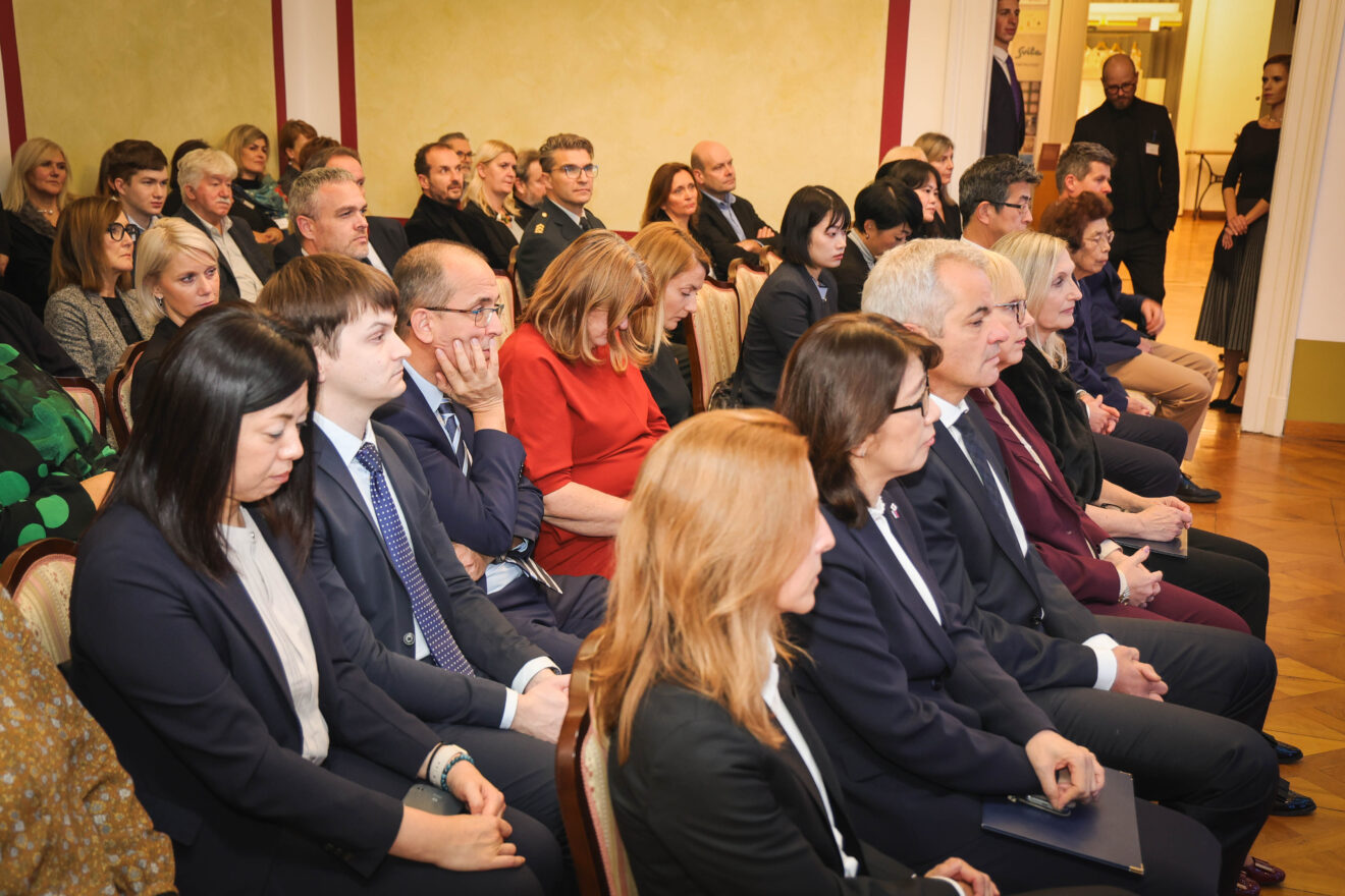 Formalno zbrano občinstvo, ki sedi v vrstah, oblečeno v poslovno opravo.