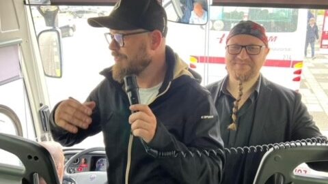 Moški z mikrofonom v črni kapi in jakni, ki vodi pogovor na sprednjem sedežu avtobusa, ob njem stoji še en moški.